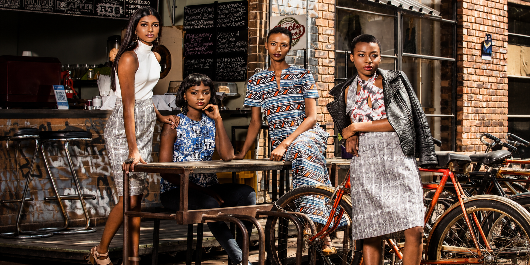Four women posing together in a cafe setting with brick walls, chalkboard menus, and bicycles in the background and all wearing KISUA Fashion brand.
