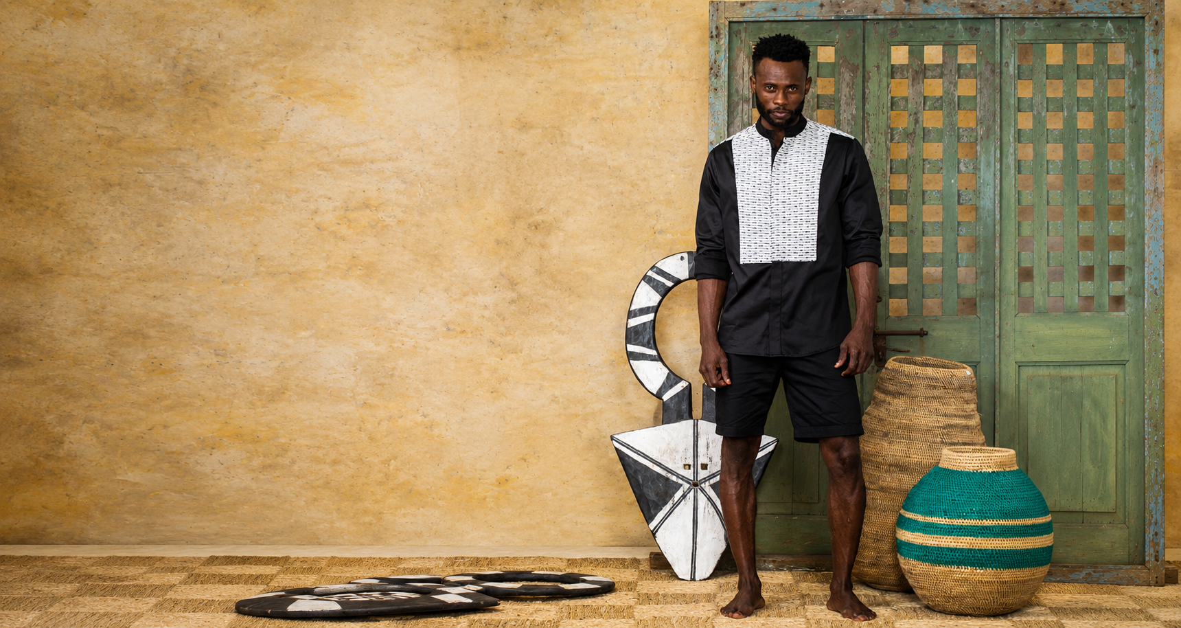 A man stands barefoot in a rustic interior wearing a black traditional outfit from KISUA Fashion, with a white patterned chest panel. 